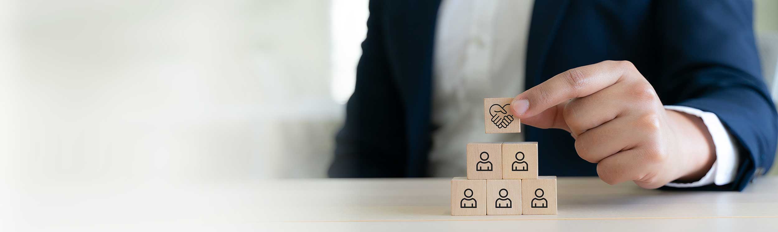 Hand platziert einen Baustein mit Handschlag-Symbol auf ein Team aus Holzklötzen als Sinnbild für die Verbindung von Business und Psychologie.